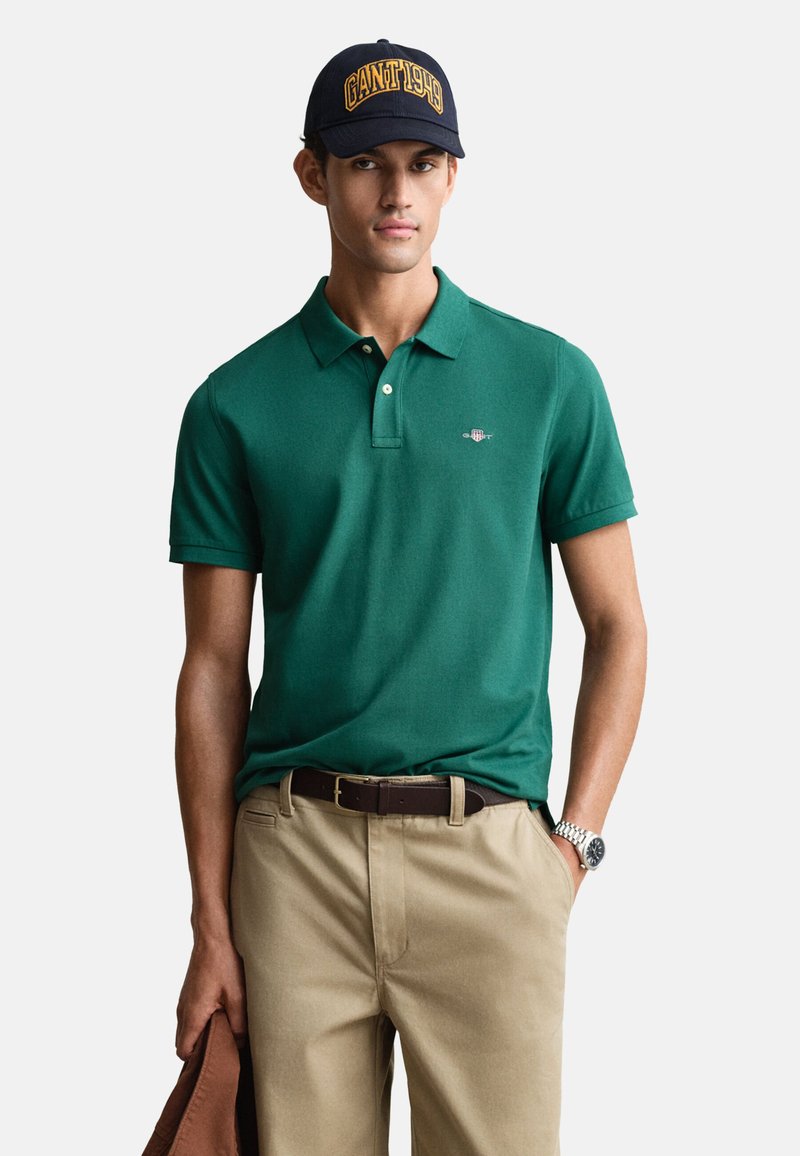 Polo vert à col court, pantalon beige, ceinture marron et casquette foncée avec l'inscription "GANT 1919" en jaune. Le modèle pose de manière neutre.
