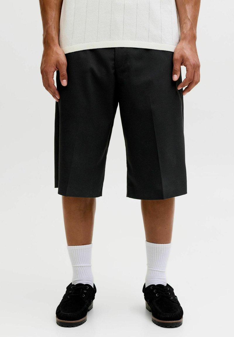 Homme portant un short noir sur mesure jusqu'aux genoux, des chaussettes blanches, des chaussures noires à lacets et un pull sans manches blanc.