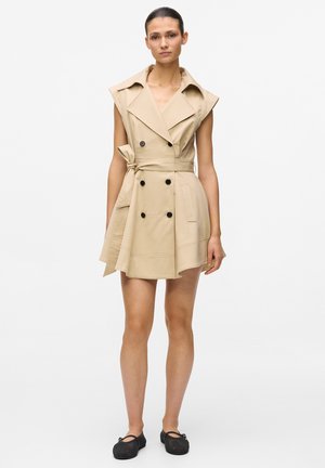 Kvinne som står iført ermeløs beige dobbelspent trenchcoat-kjole med belte og svarte flate sko mot hvit bakgrunn.