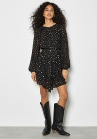 Schwarzes Kleid mit langen Ärmeln und einem Rüschensaum, verziert mit goldenen Sternmustern. Kombiniert mit schwarzen Cowboystiefeln.