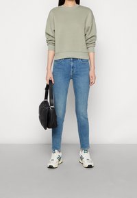 Persona con sudadera verde claro, jeans azules, zapatillas blancas con rayas verdes, sosteniendo un bolso negro acolchado de hombro.