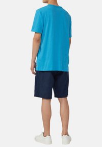 Blauwe katoenen T-shirt met korte mouwen, gedragen met marineblauwe shorts en witte sneakers. De stof heeft een lichte textuur en de pasvorm is ontspannen.