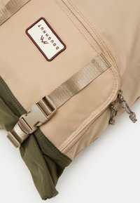 Zaino marrone con fondo verde, dotato di tasca frontale con cerniera, etichetta con marchio e spallacci in nylon con chiusura a fibbia laterale.