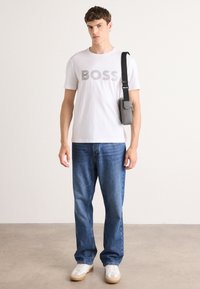 Witte crewneck t-shirt met "BOSS" in grijze letters, gecombineerd met blauwe jeans en witte sneakers met tan accenten. Kleine grijze tas op de schouder.