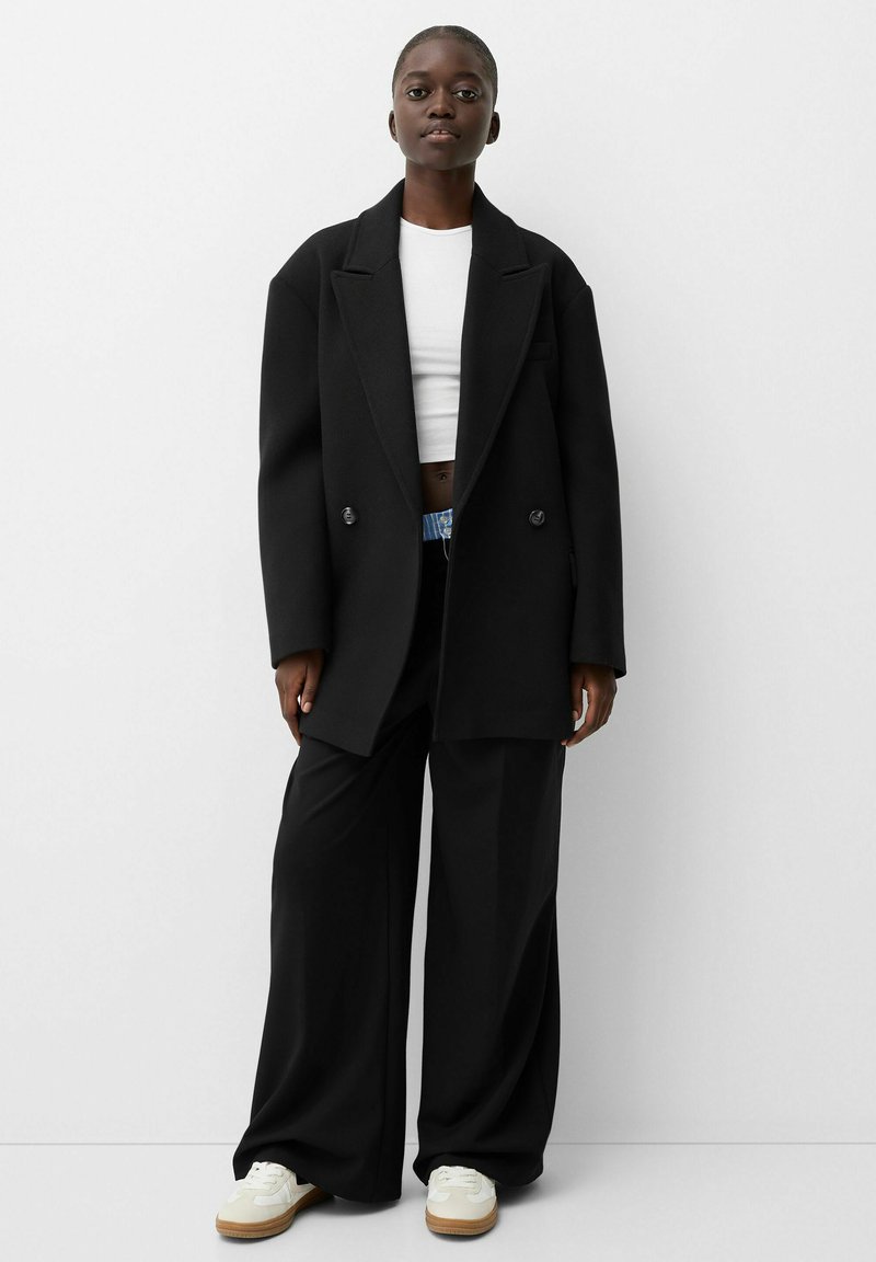 Schwarzer oversized Blazer mit doppelt geknöpften Knöpfen, kombiniert mit einem weißen Cropped-Top. Lockere schwarze Hosen und helle Sneakers mit einer Gummisohle.