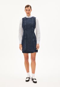 Robe en denim évasée avec des coutures visibles, couleur bleu foncé, sans manches et décolleté rond. Associée avec une chemise blanche à col et des chaussures noires.