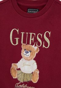 Bordeaux sweatshirt med en broderet bjørn i en hvid top og guld nederdel, med "GUESS" og "California" tekst i dekorative skrifttyper.