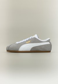 Γκρι και λευκό αθλητικό παπούτσι Puma με λευκά κορδόνια, καουτσούκ σόλα και χρυσό λογότυπο "Puma Tackle" στη πλευρά, εμφανιζόμενο σε προφίλ σε ουδέτερο φόντο.