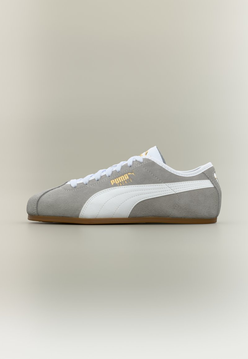 Γκρι και λευκό αθλητικό παπούτσι Puma με λευκά κορδόνια, καουτσούκ σόλα και χρυσό λογότυπο "Puma Tackle" στη πλευρά, εμφανιζόμενο σε προφίλ σε ουδέτερο φόντ ο.