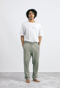 Hvid kortærmet t-shirt med "TOMMY HILFIGER" logo, parret med lysegrønne bukser med snor, begge i en afslappet pasform, på en person uden sko.