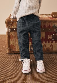 Donkere geruite broek met een slanke pasvorm en elastische tailleband, gecombineerd met witte sneakers met rode accenten, geplaatst naast een vintage koffer.