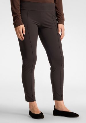 Personne portant un pantalon marron foncé ajusté et des chaussures plates noires, debout devant un fond clair uni.