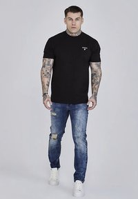 Svart bomulls-t-shirt med liten logotyp på bröstet, kombinerad med blå slitna jeans och vita sneakers. Modellen står vänd framåt.