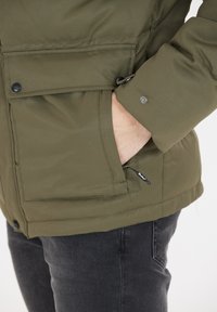 Oliven grøn isoleret jakke med trykknapper, lynlås i sidelomme og en blød tekstur. Har en justerbar bund og manchetter.