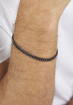 Close-up of a hairy wrist wearing a thin black metal chain bracelet against a white fabric background.