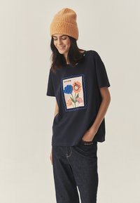 Tumesinine T-särk lillemustriga, millel on värvikas karu ja lillede tikand, kombineeritud tume teksapükste ja oranži kootud mütsiga.