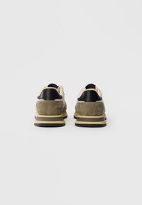 Beige suede joggesko med svarte skinnakenter, med en polstret såle, rund form og kontrasterende gul trim på mellomsålen.