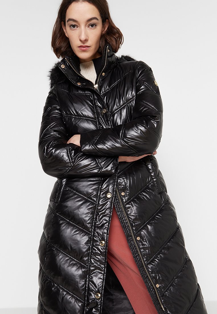 Donna che indossa un lungo cappotto imbottito nero lucido con cappuccio rifinito in pelliccia, un maglione bianco e pantaloni color ruggine, in piedi con le braccia incrociate.