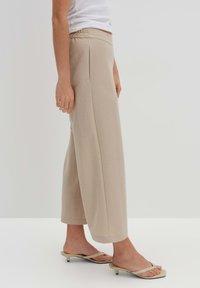 Beige Hosen mit weitem Bein und elastischem Bund, mit einer glatten Textur und lockerer Passform. Kombiniert mit hellen Rutsch-Sandalen.