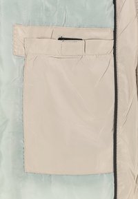 Lichte jas met een zachte, lichtblauwe binnenkant en een beige buitenzak met klepsluiting en zwarte accentstiksels.