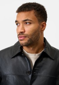 Schwarze Lederjacke mit kragenförmigem Design, ausgestattet mit einem Knopfverschluss und einer glatten Textur. Trägt sie über einem hellbeigen Pullover.