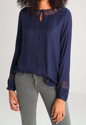 Blusa navy con scollatura rotonda, dettaglio in pizzo al collo e ai polsini, maniche lunghe e vestibilità rilassata, abbinata a jeans grigi.