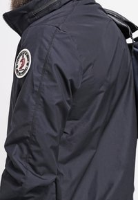 Marinblå lättviktsjacka med dragkedja vid kragen, texturerad tyg och en rund patchdetalj på vänster ärm. Har en stilren design.