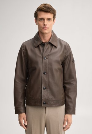 Jonge man met een bruine leren jas met knopen over beige broek, staand tegen een effen lichte achtergrond.