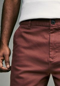 Chinos marron avec une texture lisse, présentant une fermeture à bouton, des poches latérales et une coupe ajustée, associés à une chemise blanche.