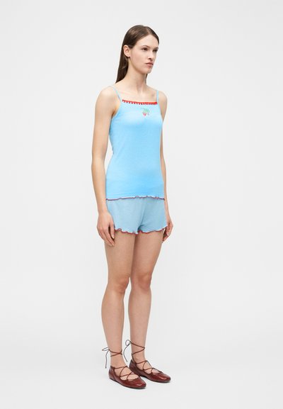 Γυναίκα που στέκεται με μπλε σορτς και τοπ με λεπτές τιράντες, κόκκινο τελείωμα και κέντημα φράουλας, φορώντας καφέ δερμάτινα παπούτσια με κορδόνια.