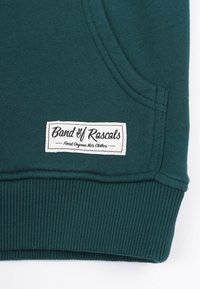 Mörkgrön sweatshirt med en mjuk textur, med en vit etikett som visar "Band of Rascals" och ribbade muddar vid fållen.