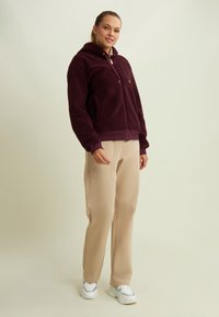 Giacca foderata bordeaux con cappuccio, dotata di tasche frontali e polsini elastici, abbinata a pantaloni beige e sneakers bianche con accenti grigi.