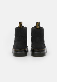 Botines de ante negro con una superficie texturizada, que cuentan con una suela gruesa negra y lengüetas amarillas. El diseño incluye costuras en contraste.