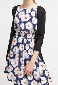 Abito floreale con sfondo navy, caratterizzato da grandi fiori bianchi con centri rossi. Cardigan nero, vita aderente e gonna al ginocchio.
