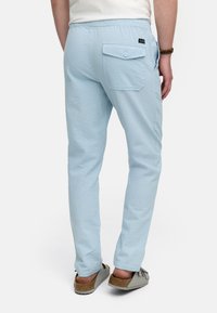 Hellblaue gewebte Hose mit strukturierter Oberfläche, slim fit, elastischem Bund und einer einzigen Knopftasche auf der Rückseite. Trägt bequeme Schuhe.