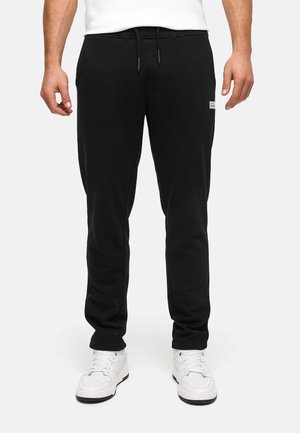 Mann trägt schwarze Sweatpants mit Kordelzugbund und weiße Sneakers, steht vor einem schlichten hellen Hintergrund.