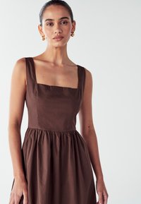 Rjava brezrokavna obleka s kvadratnim izrezom, zategnjeno pasom in razširjenim krilom. Narejena iz gladkega, lahkega materiala. Zlate uhane v obliki obroča.