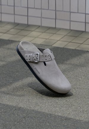 Sabot en daim gris clair à enfiler avec bride réglable à boucle et semelle noire, reposant sur un bitume gris près d’un mur carrelé.