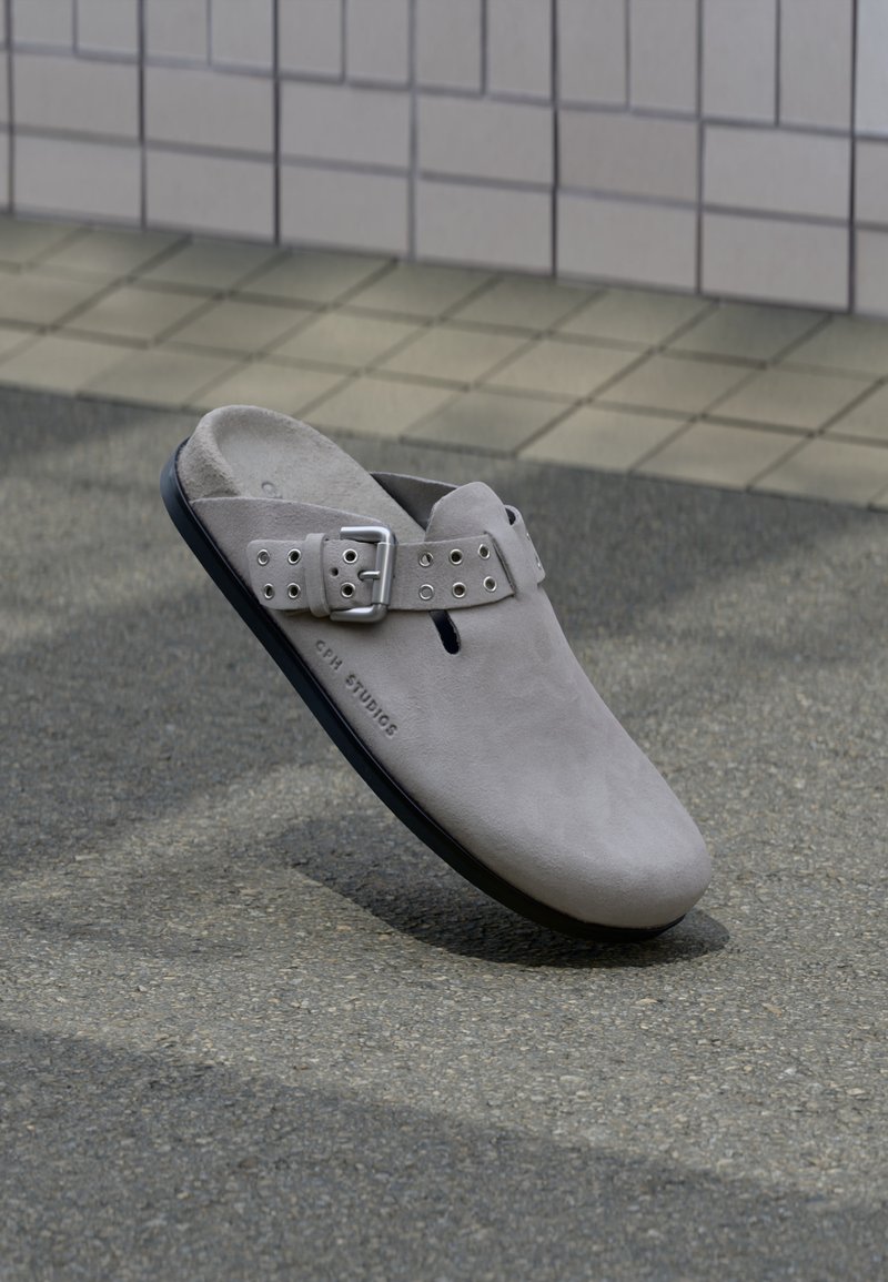 Ανοιχτό γκρι μοκασίνι από σουέτ με ελαστικό λουράκι με αγκράφα και μαύρη σόλα, ακουμπώντας σε γκρι άσφαλτο κοντά σε πλακιδωτό τοίχο.
