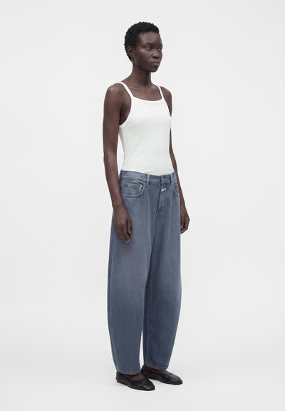 Femme portant un débardeur blanc, un jean bleu à jambes larges et des ballerines noires, debout devant un fond clair uni.