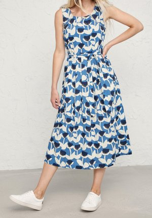 Kobieta w niebiesko-białej kwiecistej sukience midi bez rękawów z wiązaniem w talii, w połączeniu z białymi trampkami, stoi przy teksturowanej ścianie.