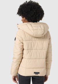 Beige puffer jacket with a hood, quilted texture, adjustable drawstring, and a logo patch on the left sleeve. Features side pockets.