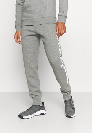 Šedé bavlněné tepláky s elastickým pasem, s výrazným bílým logem po celé levé nohavici. Kombinované se šedými sportovními botami.