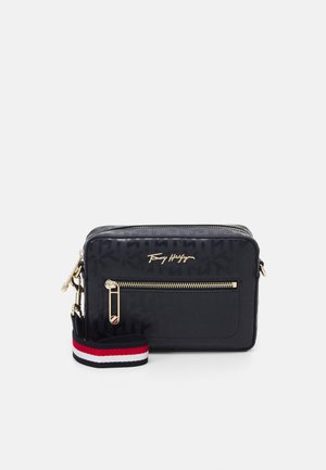 Kompaktowa czarna torba na ramię Tommy Hilfiger z przednią kieszenią na zamek, złotym logo i suwakiem oraz odpinanym paskiem w czerwono-biało-granatowe paski.