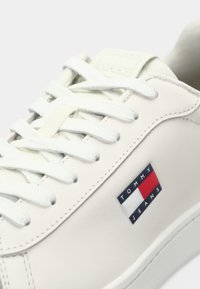 Sneaker din piele albă cu șireturi albe, având o patch rectangulară în culorile albastru, roșu și alb, inscripționată cu "TOMMY JEANS" pe lateral.