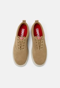 Beiga mockaskor med rund tå, snörningsdesign och röd innersula med "CAMPER"-text. Tunn vit yttersula och dragflik vid hälen.