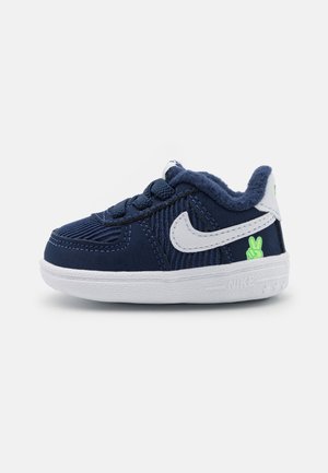 Marineblå småbørns sneaker med hvid Nike-swoosh, hvid sål, blødt indvendigt for og lille grøn fredstegn-detalje på hælen.