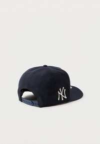 Casquette de baseball bleu marine avec une visière plate et une sangle ajustable. Présente un logo brodé blanc sur le côté gauche et un tissu texturé.