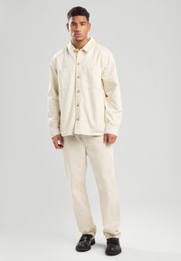 Crème kleurige overhemdjas met twee voorzakken en houten knopen, gecombineerd met bijpassende crème rechte broek en zwarte schoenen.