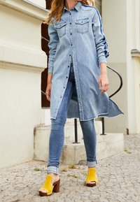 Jeansskjortklänning med långa ärmar, knappar fram och bröstfickor. Bärs över blå jeans och matchas med gula sandaletter med öppen tå och träklack.